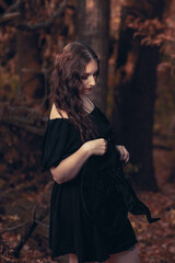 This striking photograph features a woman standing in a dark, moody forest, exuding a gothic and mysterious aura. She is dressed in an all-black ensemble, including a sheer, flowing cape.