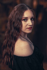 This striking photograph features a woman standing in a dark, moody forest, exuding a gothic and mysterious aura. She is dressed in an all-black ensemble, including a sheer, flowing cape.