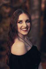 This striking photograph features a woman standing in a dark, moody forest, exuding a gothic and mysterious aura. She is dressed in an all-black ensemble, including a sheer, flowing cape.