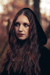 This striking photograph features a woman standing in a dark, moody forest, exuding a gothic and mysterious aura. She is dressed in an all-black ensemble, including a sheer, flowing cape.