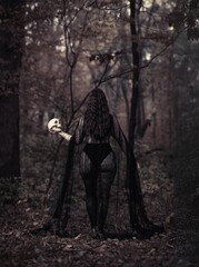 This striking photograph features a woman standing in a dark, moody forest, exuding a gothic and mysterious aura. She is dressed in an all-black ensemble, including a sheer, flowing cape.
