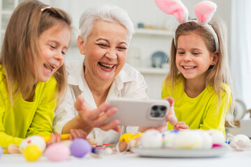 Happy Easter. Two girls sisters twins grandmother painting eggs watching video on phone enjoying time together. Happy family preparing for Easter. Good time at home. Christian festival tradition