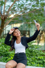 Fototapeta premium Young Asian Woman Taking Selfie with Phone Sitting Outdoors in Modern City with Greenery