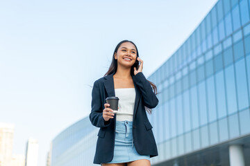 Fototapeta premium Young Asian Businesswoman Using Phone While Holding Coffee in Modern Office District Outdoors