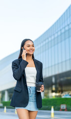 Fototapeta premium Young Asian Businesswoman Talking on Phone While Holding Coffee in Modern Office District Outdoors