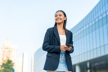 Fototapeta premium Young Asian Gen Z Businesswoman Using Phone in Casual Modern Office District Outdoors