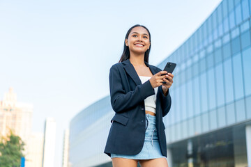 Fototapeta premium Young Asian Gen Z Businesswoman Using Phone in Casual Modern Office District Outdoors