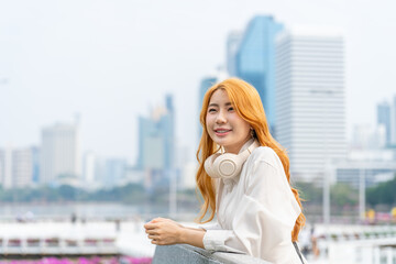 Fototapeta premium Asian woman relaxing with headphones around neck in modern cityscape. Young female enjoying outdoor break in urban environment.