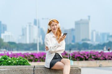 Fototapeta premium Asian woman enjoying music while using phone in city park. Relaxed young female with headphones sitting outdoors in urban landscape.