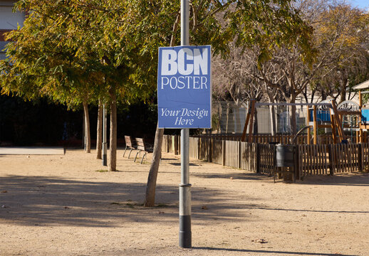 An ad poster is on a lamppost in a Barcelona children's park, surrounded by greenery and playgrounds.