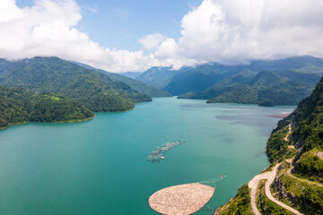 Obraz premium Aerial view of a serene mountain lake with turquoise water, surrounded by lush green hills. A fish farm floats on the calm surface, adding an element of human presence in pristine nature.