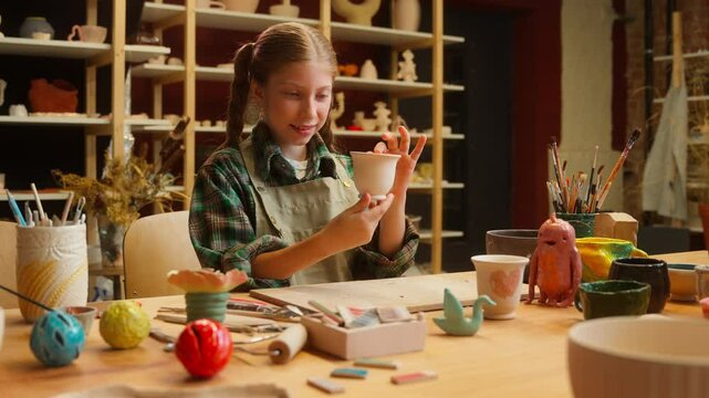 Adorable Caucasian little girl teenager child daughter school kid pottery sculptor artist potter examine inspecting clay craft vase handmade artwork crockery creative stoneware inside workshop studio