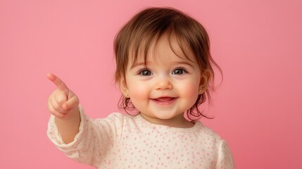 Charming baby girl with rosy cheeks enthusiastically gestures left while wearing a lovely pastel outfit on a pink background