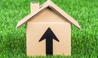 Small cardboard house with an upward arrow symbol on green grass representing growth, housing market trends, and real estate investment opportunities