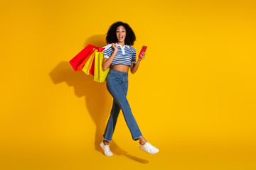 Cheerful young woman holding shopping bags and smartphone against a bright yellow background featuring casual style