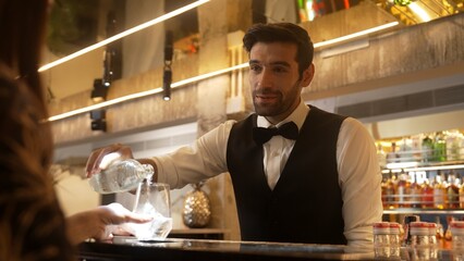 Focusing on smart bartender serving gin fizz or tonic at luxurious bar while engaging lively conversation with VIP customer at back view in lighting atmosphere at lighting warm nightclub. Vinosity.