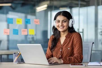 Young woman working inside office wearing headphones, programmer smiling and looking at camera, employee coding new software, developing applications.