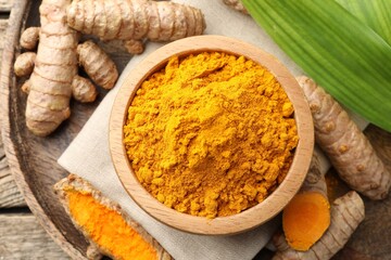 Turmeric powder in bowl, roots and leaves on wooden table, flat lay