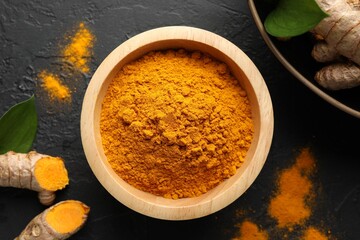 Aromatic turmeric powder in bowl, fresh roots and leaves on black table, flat lay