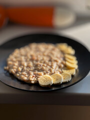 A black plate filled with creamy oatmeal, topped with banana slices and peanuts, sits on a windowsill with a blurred background.
