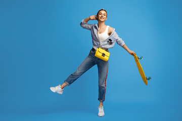 Charming young woman with yellow skateboard, soccer ball and headphones dancing on blue background