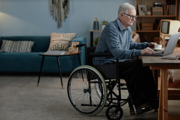 Full length shot of senior male wheelchair user typing on laptop sitting at desk communicating...