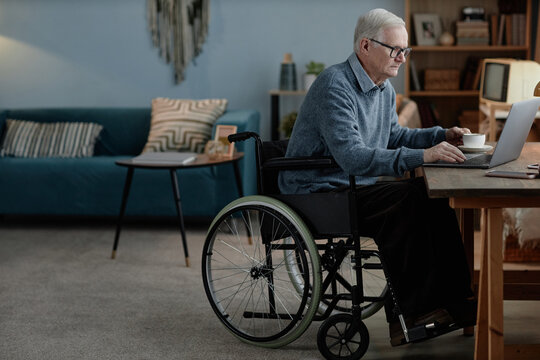 Full length shot of senior male wheelchair user typing on laptop sitting at desk communicating online independently in living room, copy space