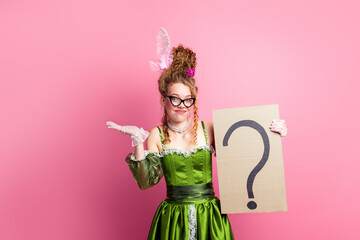 Young lady in historical green gown holding a question mark sign against pink background