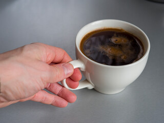 Hand gently grasps the handle of a white mug filled with dark, aromatic coffee, resting on a simple gray table, creating a cozy and inviting scene
