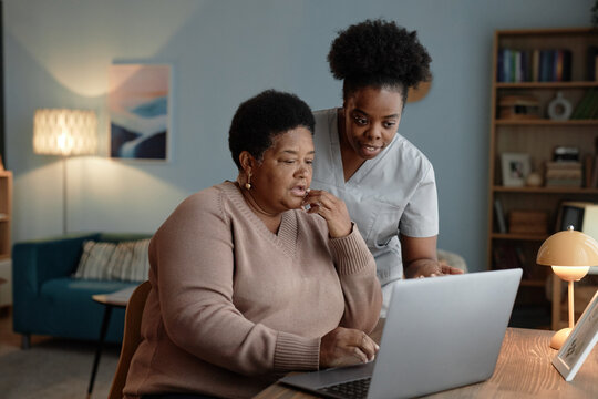 Supportive African American female social worker assisting elderly woman with laptop use explaining how to pay bills online while sitting at desk in cozy living room, copy space