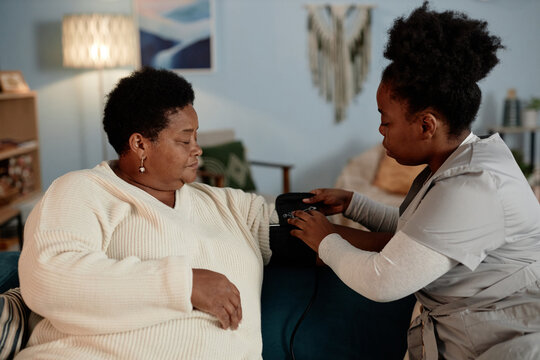 Female nurse wrapping cuff of automatic tonometer on elderly Black womans arm measuring blood pressure during health checkup visit at home