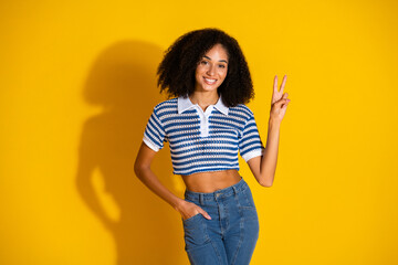 Fashionable young woman in a striped polo shirt posing with a peace gesture on a vibrant yellow...