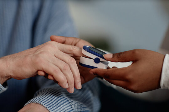 Close up on hand of elderly man putting fingertip on oximeter sensor while Black female social worker measuring blood oxygen saturation during home care visit, image in warm tones, copy space
