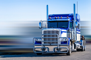 Classic bonnet American idol big rig blue and white semi truck tractor with pipe grille guard and chrome accessories standing without semi trailer on industrial parking lot with blurry background