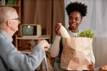 Medium portrait shot in warm tones of accommodating Black female social worker unpacking paper bag...