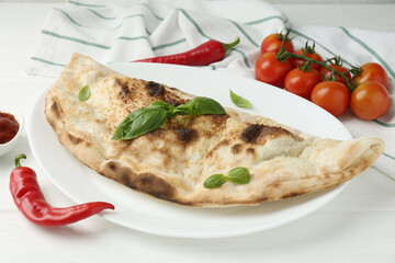 Tasty calzone, basil, chili pepper, sauce and tomatoes on white wooden table, closeup