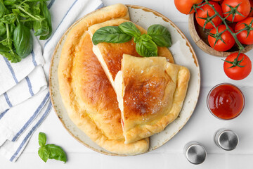 Pieces of delicious calzone pizza with mozzarella, tomatoes and basil on white tiled table, flat lay