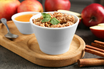 Delicious apple crisp in bowl, fresh fruits, cinnamon sticks, honey and mint on grey table, closeup