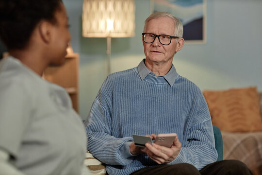Medium portrait of white haired elderly man making electronic payment online on mobile phone while social worker assisting pensioner at home