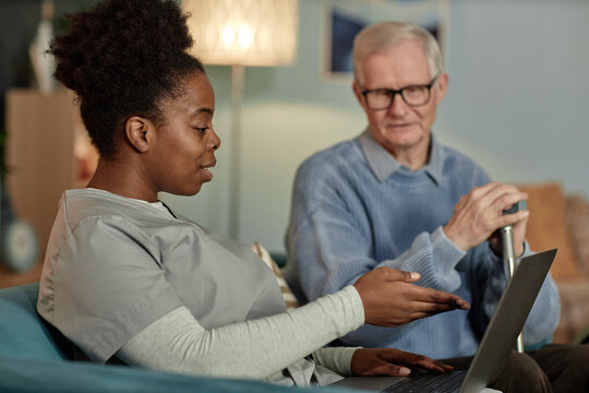 Adult African American female social worker assisting elderly man with online payment services pointing at laptop screen while sitting together on couch in living room