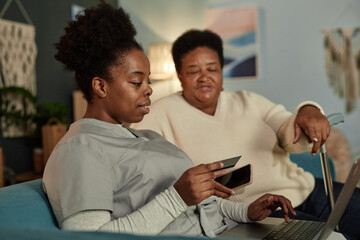 Side view of African American female social worker typing on laptop assisting elderly woman with...