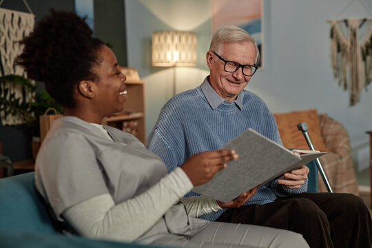 Medium shot of happy elderly man looking through photograph album with cheerful female social worker, sitting together on couch in living room during home care visit