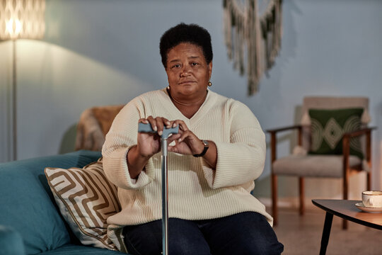 Medium portrait of calm elderly Black woman wearing beige knitted sweater sitting alone on couch holding walking cane at home, copy space