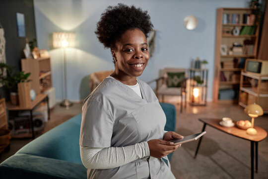 Medium portrait of African American female social worker in scrubs holding digital tablet and smiling friendly at camera captured in patients house during home visit, copy space