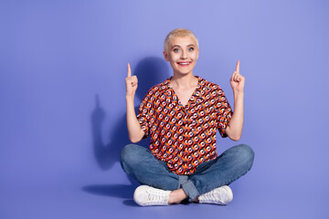 Young stylish woman smiling and pointing upward while seated cross-legged against a vibrant purple...
