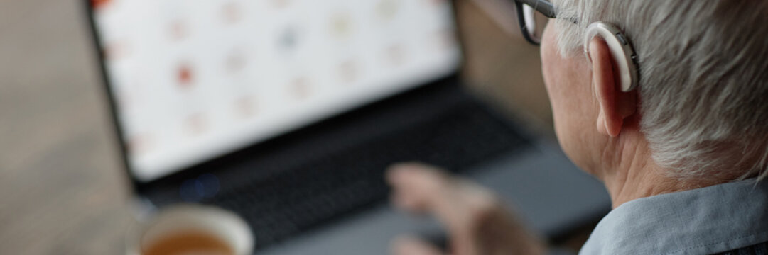 Website header shot of elderly man wearing hearing aid behind ear using laptop at home with help of assistive technology, copy space