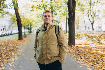 Blonde young happy 30s man wearing backpack standing on city autumn park street. Urban lifestyle concept. High quality photo