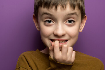 Close-up portrait of a young boy biting his nails, showing a nervous expression. The child wears a brown sweater against a vibrant purple background. Concept of anxiety and habits.