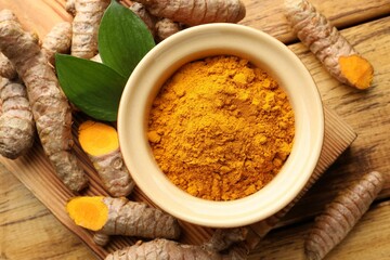 Turmeric powder in bowl, roots and leaves on wooden table, flat lay
