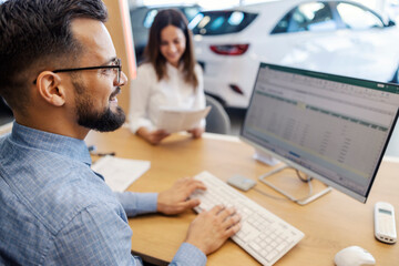 Smiling car dealer sitting at car salon and typing documents on computer while buyer reading...